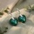 Srebrne kolczyki z kryształami serca Emerald w zbliżeniu. Zdjęcie eksponuje szlif i kontrast koloru z jasną oprawą.